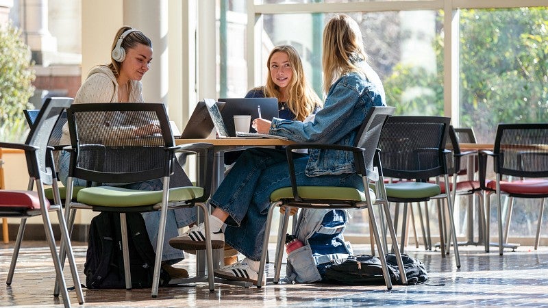 Three students sitting at a table in the Lillis atrium working on laptops and talking to each other.