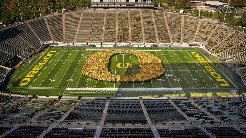 An aerial photo of a crowd of thousands of students standing together in an O shape on the field at Autzen Stadium.
