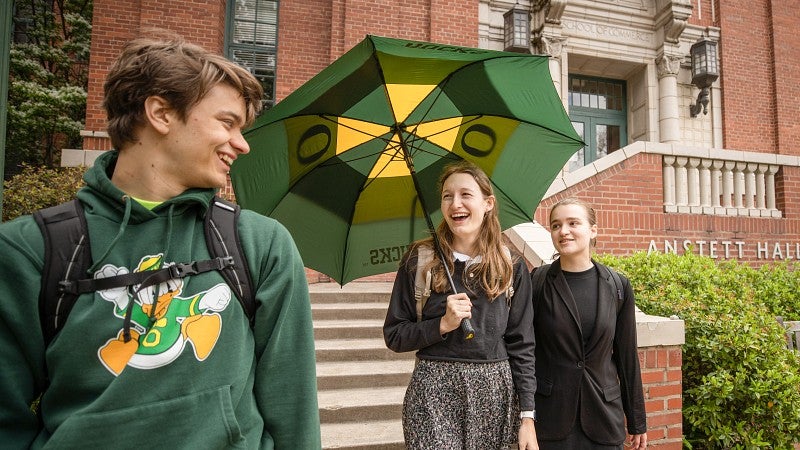 Three students walking in front of Anstett Hall laughing and talking to each other. One is holding a large opened Oregon Ducks umbrella.