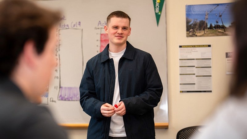 One student is standing with their back to a whiteboard, facing another student in the foreground sitting at a table.