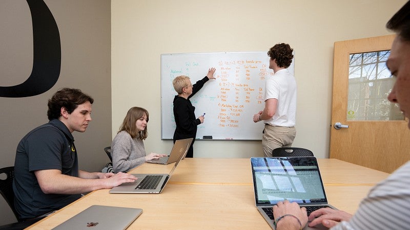 A professor and a student are standing at a whiteboard mid-conversation and pointing at accounting figures. Three other students are sitting at a table on their laptops.