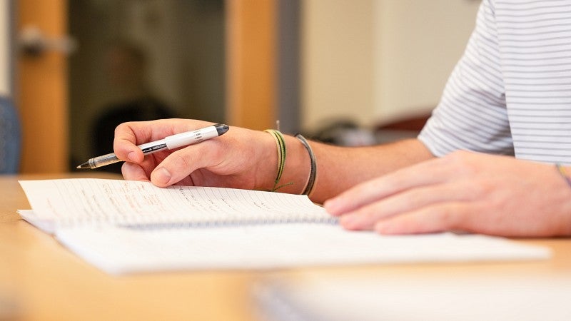 A close-up of a set of hands holding a pen and thumbing through a hand-written notebook.