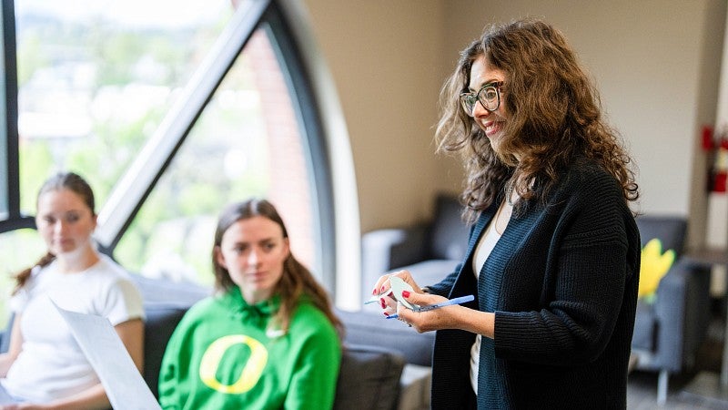Two students sitting in chairs glancing at a professor standing next to them, smiling.