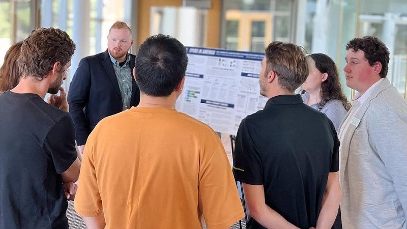 A group of students standing around and listening to another student's presentation on an easel.