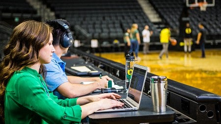 Two students sitting on the sidelines of a basketball court using laptops