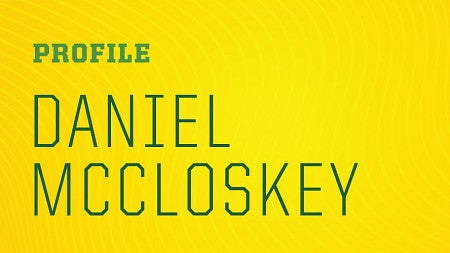 Photo of Daniel McCloskey in cap and gown through the Oregon "O" with his hands to the left of the words "Profile" Daniel McCloskey" in large letters on a yellow background