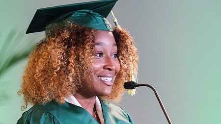 Sports Product Management graduate Kiara Franklin, wearing a graduation cap and gown, speaks at the podium during the SPM online program's commencement ceremony