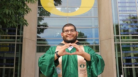 David Olvera, wearing commencement regalia and making the "O" symbol with his hands, stands beneath the giant "O" on the south face of the Lillis Business Complex