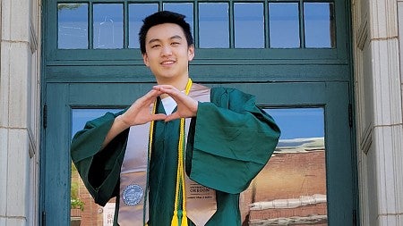 Ethan Wong, wearing commencement regalia and standing outside an entrance to the Lillis Business Complex, makes the "O" shape with his hands