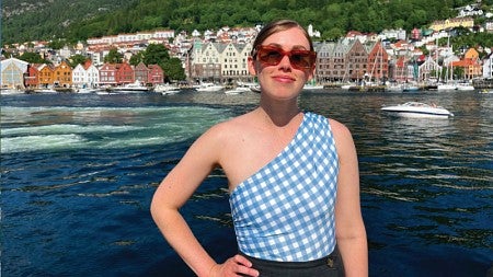 Tess Meyer pictured on a sunny day in Sweden. Behind her is a waterfront scene including docked boats and a town lining the banks.
