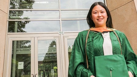 Ping Wang, wearing commencement regalia, stands outside the Lillis Business Complex underneath the giant "O" on the south face of the building