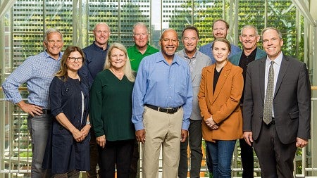 Photo of the board of advisors group from 2023 standing in two rows facing the camera while on the fourth floor landing of the Lillis Business Complex atrium, with the photovolatic cells embedded in the buildings south class wall visibile in the background.