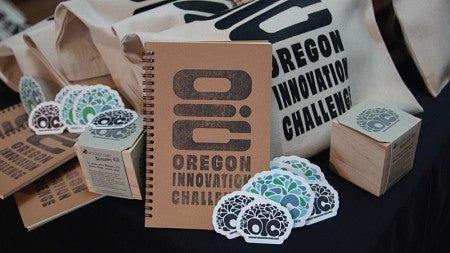 Photo of a pile of tote bags, notebooks, and stickers branded with the Oregon Innovation Challenge logo
