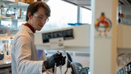 Biochemistry PhD student Justin Svendson, wearing a lab coat and goggles, works on equipment in a laboratory.