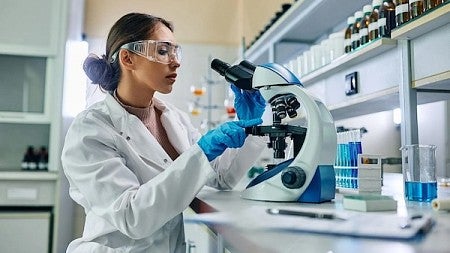 A female scientist looks into a microscope in a lab setting while wearing protective eyewear and a lab coat