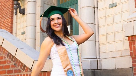 Lorena Garcia ’24 (business administration), wearing her graduation cap on her head and her cords and sash around her neck, poses for a photo in front of Anstett Hall. She has her right hand on her commencement gown, which is draped over the bannister of the stairs she's leaning against, and is lifting her left arm to touch her cap with her other hand.