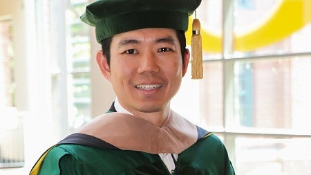 Woocheol Kim, PhD ’24 (marketing), wearing his commencement regalia, stands in the atrium of the Lillis Business Complex on a sunny day.