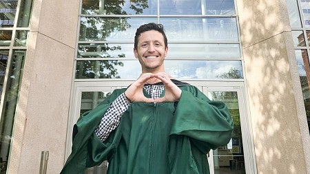 Brodi Sabiston, MBA ’24, MS ’24 (finance), standing outside the south entrance to the Lillis Business Complex while wearing his commencement regalia, makes the "O" symbol with his hands.
