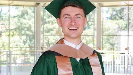 James Schulte, MBA ’24, dressed in commencement regalia, stands in the atrium of the Lillis Business Complex on a sunny day.