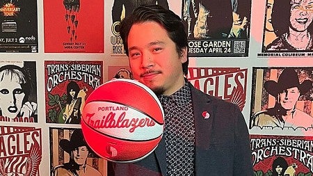 Lundquist College alumnus Ryan Horiuchi, standing in front of wall collaged with music posters, holds a Portland Trailblazers basketball in front of him.