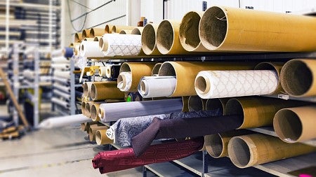 Rolls of fabric peek out of cardboard tubes stacked on shelves on a factory floor.