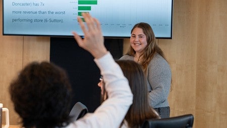 A student raises their hand while another student makes a presentation at the front of a classroom