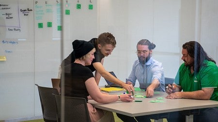 Four students work together at a table; behind them is a whiteboard covered in notes