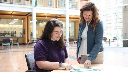 Emily Severeid (left) and Kim Thompson (right) look at some documents together at a table in the atrium of the Lillis Business Complex.