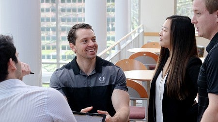 Two students sitting at a table in the Lillis atrium speak to two others standing beside them