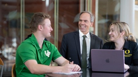 Lundquist College dean Bruce Blonigen stands between two students sitting at a high table as all smile and chat.