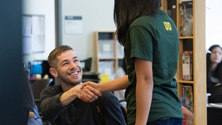 A student sitting down shakes the hand of another standing beside him