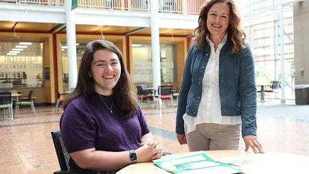 Emily Severeid (left) and Kim Thompson (right) smile as they review a document together in the atrium of the Lillis Business Complex