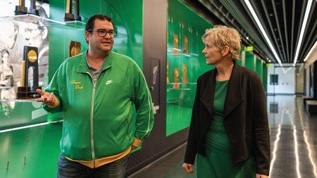 Yoav Dubinsky (left) and T. Bettina Cornwell (right) talk as they walk down a hallway filled with UO's athletic trophies