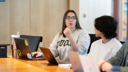 Two students sitting at a large table engaged in discussion with their laptops open.