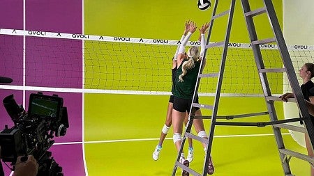 Two volleyball players on either side of the net leap up to reach the ball as a photographer films