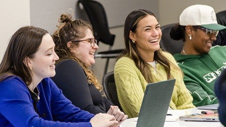 Four students sitting in a row in a classroom looking forward and smiling or laughing as a reaction to the professor.