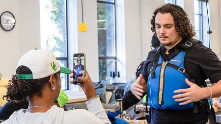 An SPM student tries on a vest design while another student takes a photo with a phone