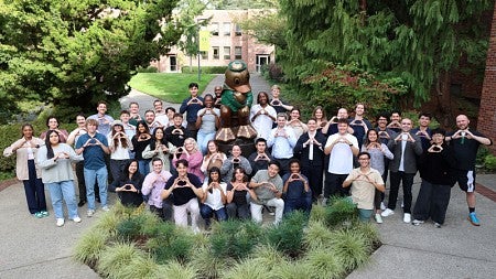 The Class of 2027 pose for a group photo outside by a Thinker-style statue of the UO Duck mascot
