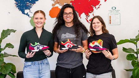 Three SPM students stand in front of a wall with a map, each holding a sneaker