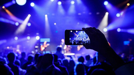 The silhouette of a person's arm and hand holding up a phone to film is seen against the bright lights of a concert on a stage