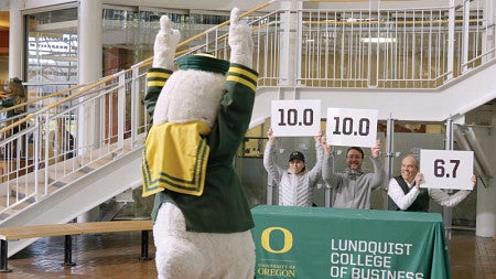 The Oregon Duck mascot holds its hands up in celebration as a judging panel of Lundquist College faculty hold up signs with numbers
