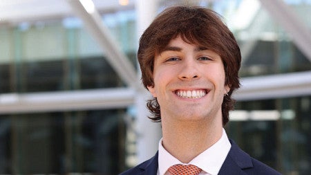 Young man with brown hair smiles in a well light room with windows behind him. Portrait is overlaid on a yellow and green background.