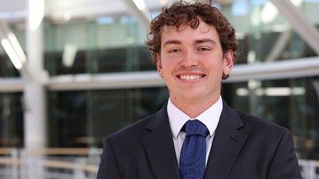 Young man with short curly hair smiles. He is wearing a dark blue suit, white shirt, and dark blue paisley tie.