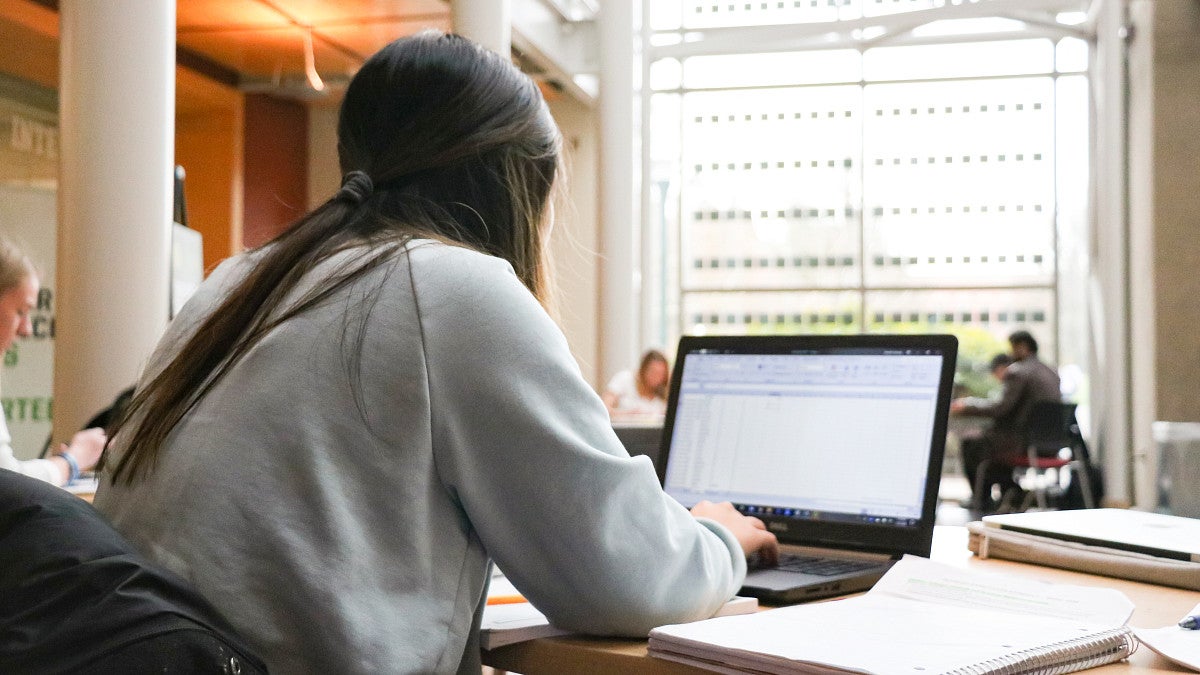 Student with their back to the camera sitting at a table working on an assignment in the Lillis Business Complex atrium.