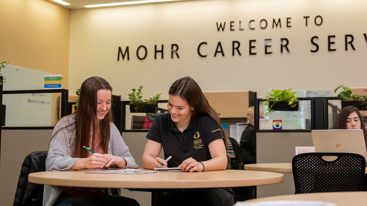 Two students sitting at a table smiling and looking at paperwork.