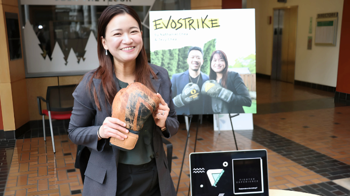 A student is holding up a prototype of her boxing glove for her startup "Evostrike" and standing in front of a poster with her project details.