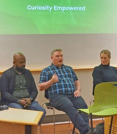 A panel of five industry representatives talk to an operations and business analytics class with the middle person in mid sentence while the four other panelist glance at him