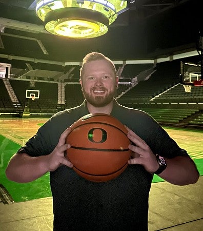 Quinn Van Horne stands courtside inside Matthew Knight Arena, smiling at the camera and holding a UO-branded basketball.