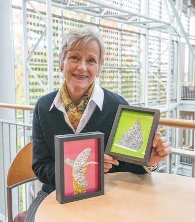 A professor is sitting at a table smiling at the camera and holding two pieces of art they created.