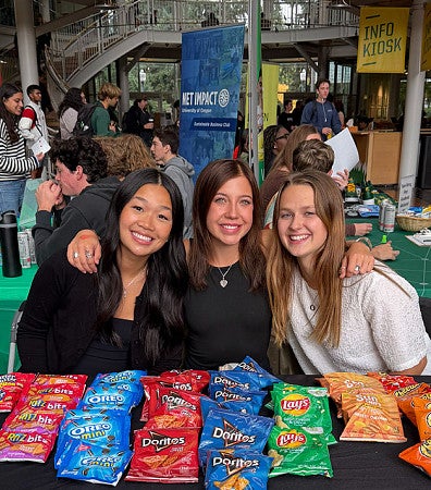 Three students sit at a table offering snacks during an event